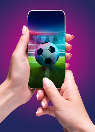 Person holding smartphone with soccer ball on stadium and filled stands in the background during open air match event.の写真素材