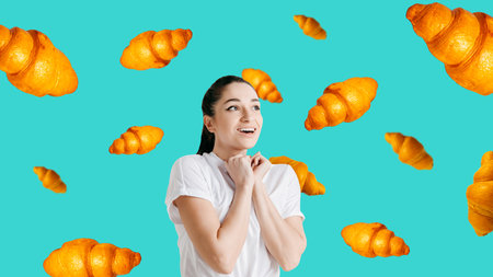 Excited young woman in white t-shirt looking up surrounded by flying croissants over vibrant turquoise background.の写真素材