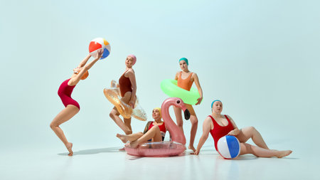 Five young women in multicolored swimsuit and caps posing with beach essentials, balls and inflatable toys against light blue backgroundの写真素材