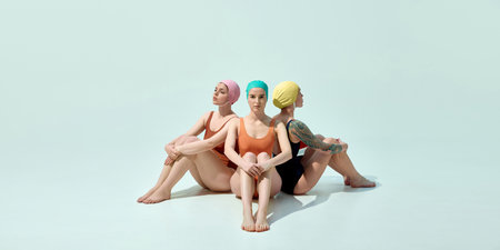Three women sitting back to back on floor in colorful swimsuits and vintage caps looking thoughtful against light blue backgroundの写真素材