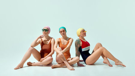 Three women in vintage swimsuits and caps sitting on floor wearing sunglasses and looking relaxed on light blue background.の写真素材
