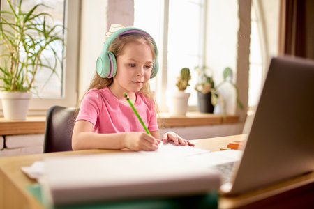 Girl in headphones writing in notebook while attentively watching laptop screen during online class at home.の写真素材