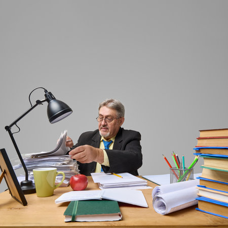 Senior teacher flipping through paper stack under desk lamp on grey background.の写真素材
