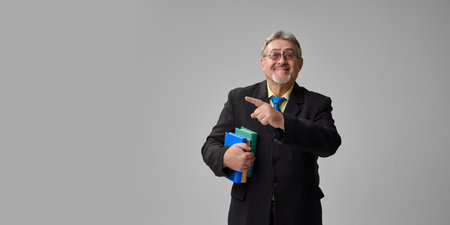 Senior teacher pointing sideways while smiling and holding books on grey background.の写真素材