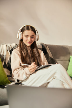 Smiling teen girl sitting on couch in headphones, attending online lesson at home, making notes on digital tablet with stylus.の写真素材