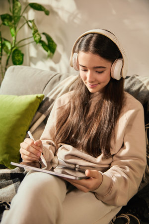 Girl smiling and sketching on digital tablet in headphones, enjoying artistic activity in cozy,...