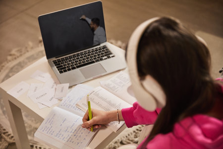 Teen girl in headphones sitting on couch, watching teacher on laptop screen and copying notes into notebook, learning words, making flashcardsの写真素材