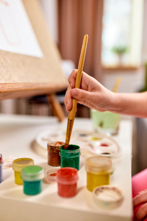 Vertical closeup of child hand dipping brush in paint jar during creative learning activity at home.の写真素材