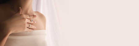 Cropped image of elegant young woman tenderly touching silver necklace with heart pendant and wearing ring, against a softly lit background.の写真素材