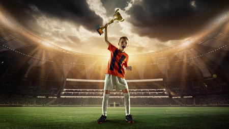 Happy and excited teen boy in orange jersey raising golden trophy with roar of victory under glowing stadium lights. 3D render of stadium.の写真素材