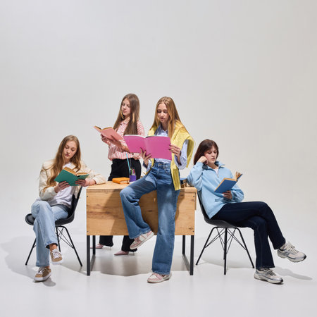 Four students in casual clothes reading books around wooden desk, showing informal collaborative homework time on grey background.の写真素材