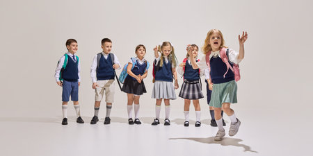 Cheerful group of first-year schoolchildren in uniform stand in line with backpacks as one girl runs...
