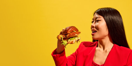 Side view of stylish young woman in red jacket preparing to bite giant burger against bright yellow background.の写真素材