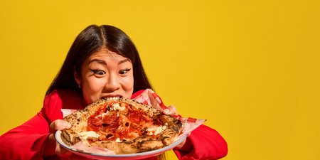 Excited Asian woman woman gripping pizza and biting edge with exaggerated emotions against bright...
