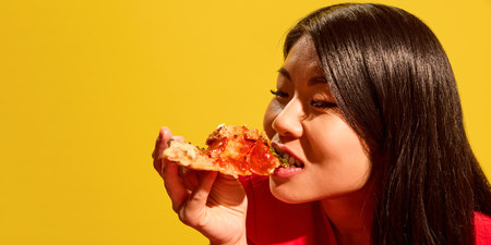 Close side profile of young Asian woman joyfully eating pizza slice with tomato sauce against bright yellow background.の写真素材