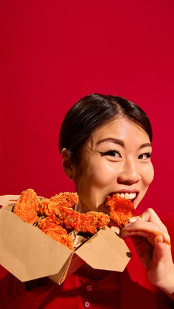 Close-up of emotional Asian woman eating delicious crispy chicken nuggets with tomato sauce from carton box against red background.の写真素材