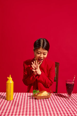 Excited woman in red suit stares at delicious hot dog on red-checkered table with mustard bottle and drink against bold red background.の写真素材