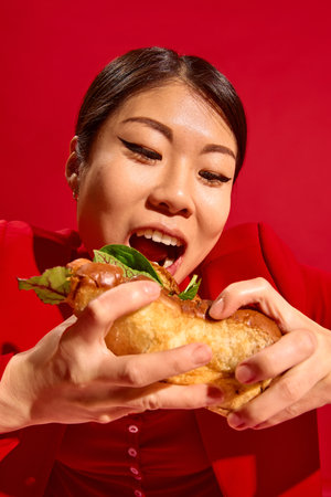 Delighted woman holds large sandwich with both hands and prepares for first bite, radiating playful emotion against red background.の写真素材