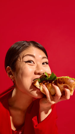 Happy Asian woman biting delicious sandwich with joy, wide-eyed and expressive face against bold red background.の写真素材