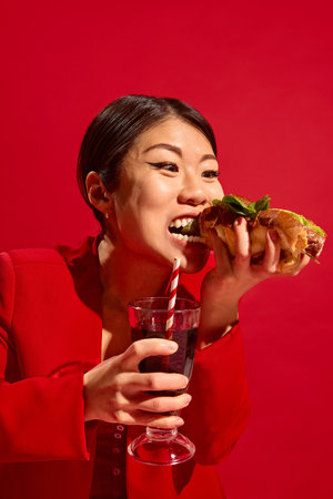 Stylish woman bites large hot dog, holding soda glass with striped straw, smiling with joy in vibrant red background.の写真素材