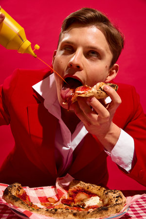 Man with delightful face pouring mustard while licking pizza slice against bold red background.の写真素材