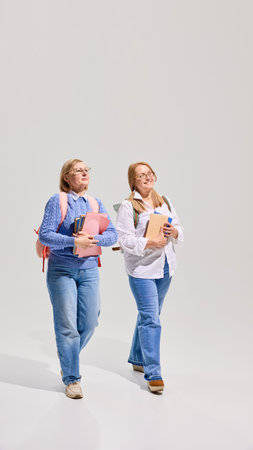 Full-length of two cheerful senior women with backpacks and books walking and talking giant white background, Elderly students.の写真素材