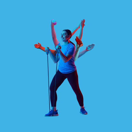 Happy sporty woman multitasking with resistance bands and dumbbells on blue background