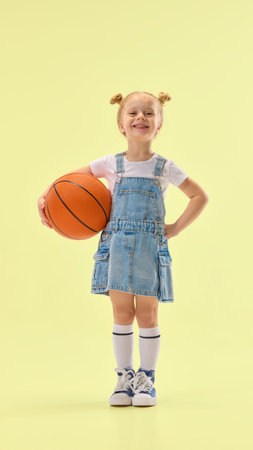 Confident girl smiling, holding basketball on hip, wearing denim overall dress and standing proud, showing positive expression on yellow background.の写真素材