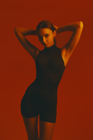 Portrait of attractive girl in black jumpsuit posing with confidence and elegance against red studio background in neon light.の写真素材