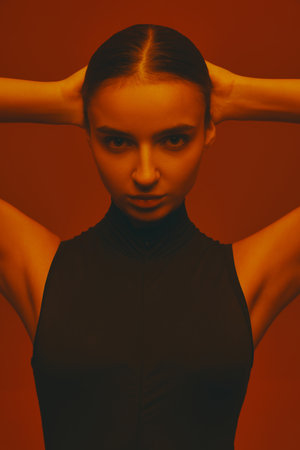 Portrait of confident and serious young wo,an in ponytail, wearing black outfit, posing with intense look against red studio background in neon light.の写真素材