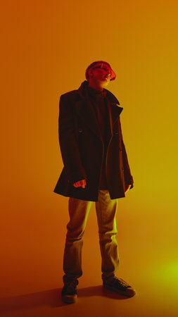Full-length portrait of stylish, confident young man in jeans, black coat and trend red sunglasses, posing against yellow background in neon lightの写真素材