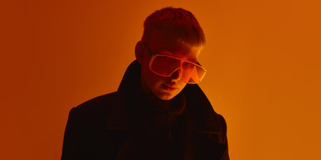 Close up of man wearing oversized orange sunglasses, looking seriously at camera under intense light against orange studio background.の写真素材
