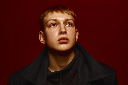 Close-up portrait of young man in dark jacket posing against rich red background, looking upwards with intense expression.の写真素材