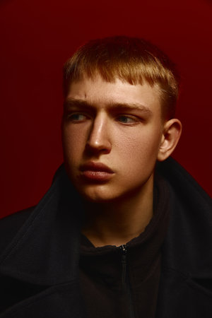 Stylish young man in black coat looks aside with serious expression under deep red studio lighting setup.の写真素材