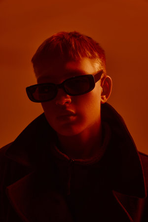 Close-up portrait of young stylish guy in trendy sunglasses and black coat, posing against red studio background in neon light.の写真素材