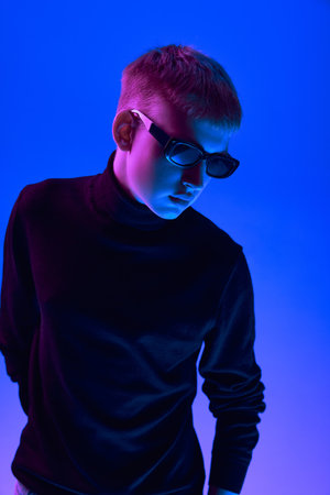 Portrait of stylish young guy in sunglasses and black turtleneck posing against blue background in neon light.の写真素材