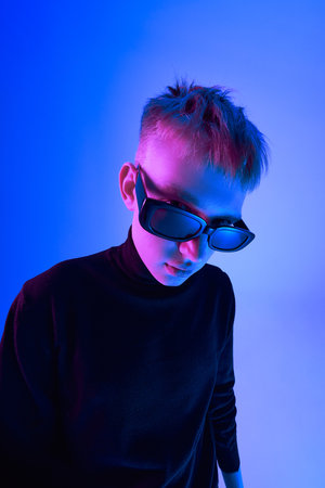 Close-up portrait of stylish young guy with short hairstyle, wearing sunglasses and black turtleneck, posing against blue background in neon lightの写真素材
