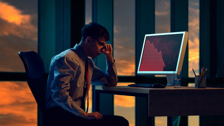 Businessman looking at downward trending graph on computer screen, with his hand on his forehead in office setting late at nightの写真素材