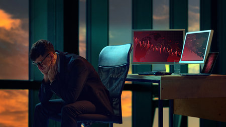 Businessman in suit sitting in office, two computer screens displaying downward trending graphs.の写真素材
