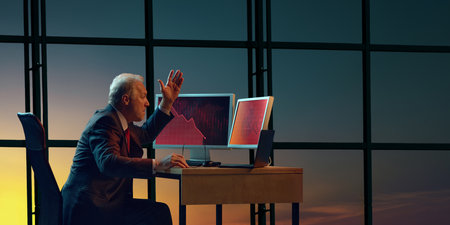 Businessman sitting at desk, raising hand in frustration while looking at two computer screens displaying red downward trending graphs.の写真素材