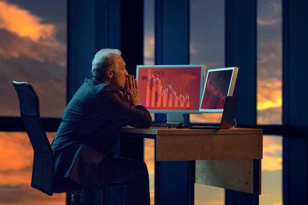Elderly businessman in sitting late in office, looking at two computer screens displaying red graphs...