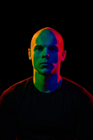 Portrait of young, handsome, bald man looking straight towards a camera with serious expression against black background in neon lightsの写真素材