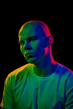 Front-angled portrait of young bald man in white t-shirt, looking towards the camera with neutral expression on dark background in multi-colored neonの写真素材