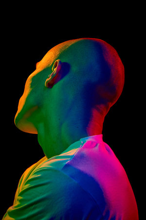 Back-angled portrait of young bald man in white t-shirt, looking upwards and to the left on a dark background in multi-colored neon light.の写真素材
