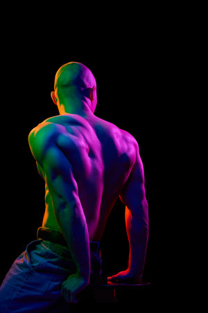 Back view of young bald man, leaning on his hands, posing shirtless, showing muscular body on dark background in multi-colored neon light.の写真素材