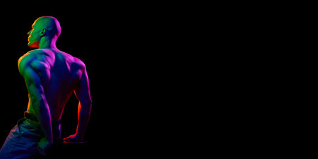 Side-angled view of young bald man, shirtless, demonstrating strong, muscular body against dark background in multi-colored neon lightの写真素材