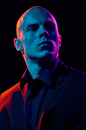 Close-up portrait of handsome bald man in dark shirt and jacket,posing confidently on dark background in multi-colored neon lightの写真素材