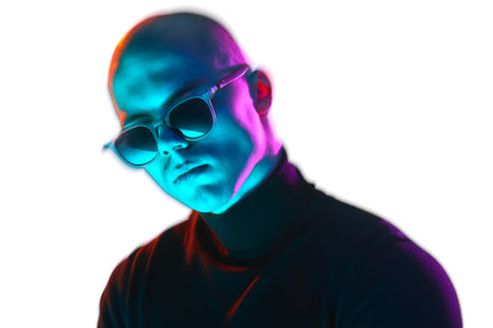 Portrait of handsome young bald man in a black turtleneck and sunglasses, confidently posing on white background in blue and pink neon light.の写真素材