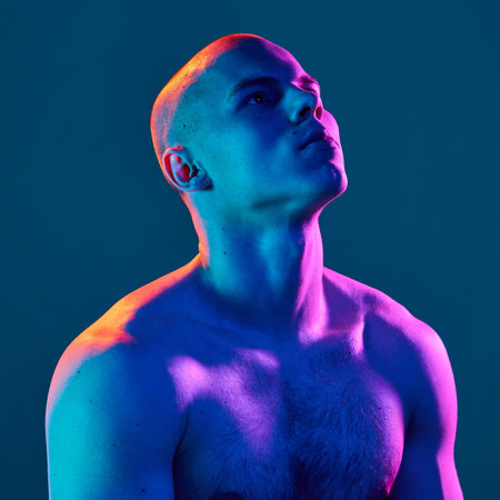 Handsome young bald man with muscular body, posing shirtless, looking upwards with neutral expression on blue background in neon lightの写真素材