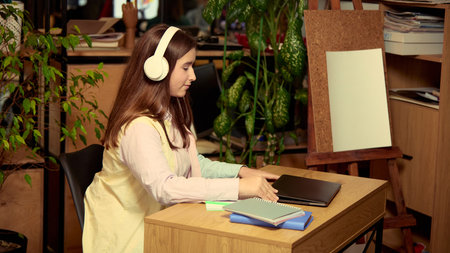 Teenage girl wearing headphones and sitting at desk with laptop and notebooks. Online learning